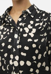 Chemise noir et blanc à motifs à pois, avec un col et des détails de boutons, fabriquée dans un tissu léger et froissé.