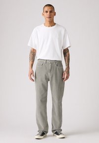Jonge man met tatoeages, gekleed in een wit oversized t-shirt, grijze ribfluweel broek en zwarte sneakers, staande tegen een effen achtergrond.