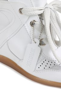 Λευκό δερμάτινο αθλητικό παπούτσι με χοντρές λευκές κορδόνια, καφέ σόλα, διάτρητη περιοχή στα δάχτυλα και ένα λευκό ετικέτα με την ένδειξη "BADONX" προσαρτημένη στο οφθαλμός.