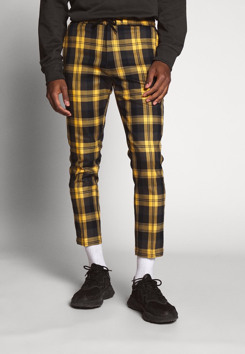 Pantalones de cuadros negros y amarillos con cintura elástica y cordón, con piernas ajustadas. Combinados con zapatillas negras y calcetines blancos.