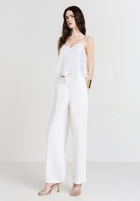 White sequined crop top with thin straps paired with high-waisted, flowy white trousers and metallic heeled slingback shoes. Gold cuff bracelet.