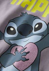 Grafisk T-skjorte med en blå tegneseriefigur med et rosa glitrende hjerte, satt mot en lys lilla bakgrunn og fet gul tekst.