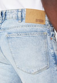 Los jeans de mezclilla azul claro tienen un corte recto, costuras visibles y un parche de cuero marrón con "DRYKORN" estampado en la cinturilla.