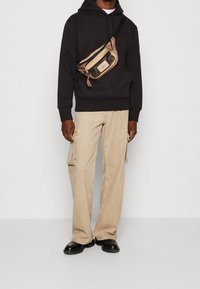 Man med svart hoodie, beige cargobyxor, svarta skor och en beige och svart crossbody-väska som står mot en enkel vit bakgrund.