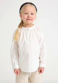 Cămașă albă cu mâneci lungi, cu decolteu rotund, nasturi și margine ondulată. Material moale din bumbac, design simplu și croială lejeră.