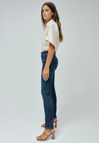 Blusa beige claro, jeans ajustados de color azul oscuro y sandalias de tacón alto en tono marrón. La ropa presenta una tela suave y un diseño clásico de mezclilla.