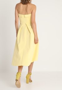 Vestido midi amarillo sin tirantes con un corsé ajustado y falda plisada. Presenta una cremallera oculta y una tela suave y texturizada. Combinado con sandalias de tacón amarillas.