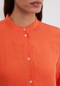 Oranžinė lino bluzė su mandarinų apykakle, turinti baltus mygtukų užsegimus ir tekstūruotą paviršių, matoma nuo kaklo linijos žemyn.