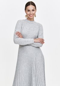 Frau mit dunklem Haar in einem grauen, langärmeligen, gerippten Kleid lächelt mit verschränkten Armen und trägt große goldene blumenförmige Ohrringe.