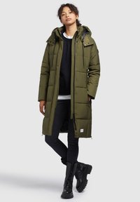 Olive green long puffer coat with a hood, zippered front, and adjustable drawstrings. Paired with black jeans and chunky lace-up boots.