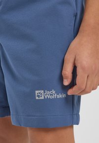 Pantalones cortos azules de material suave presentan un corte recto y un dobladillo en la parte inferior, con un sutil logo blanco de Jack Wolfskin cerca del bolsillo.