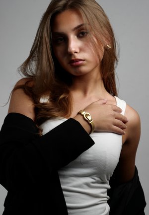 Un bracelet doré avec un grand fermoir est porté sur un bras nu, accompagné d'un débardeur blanc et d'un blazer noir drapé sur l'épaule.