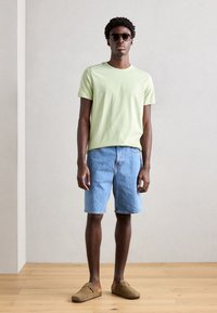 Grønn bomulls t-skjorte, lyseblå denimshorts og brune semskede slip-on sko. Modell iført solbriller, stående mot en nøytral bakgrunn.