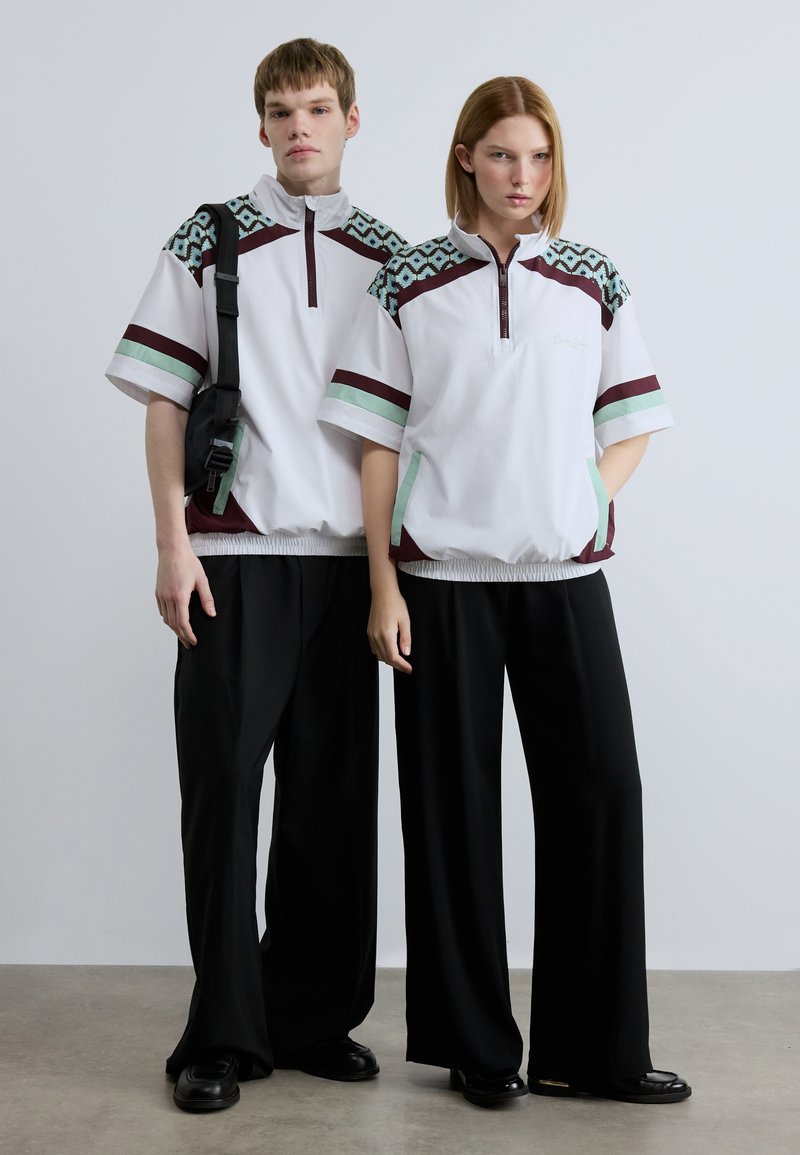 Λευκά μπουφάν με φερμουάρ μέχρι τη μέση και γεωμετρικά σχέδια σε τυρκουάζ και μπορντό, συνδυασμένα με μαύρο παντελόνι φαρδιάς γραμμής. Το ύφασμα φαίνεται λείο και ελαφρύ.