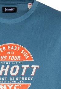 Camiseta de algodón azul con cuello redondo que presenta un gráfico de estilo vintage en naranja y blanco que dice "Schott" y "East Side Tour."