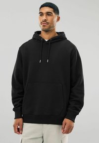Mann in einem schwarzen Kapuzenpullover mit Kordelzügen und Kängurutasche, kombiniert mit hellen Hosen, steht vor einem schlichten Hintergrund.