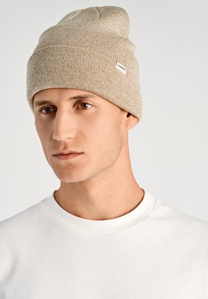 Jasnobrązowa dziergana czapka z bezszwowym wzorem i małą białą metką, noszona przez osobę w prostym białym t-shircie z okrągłym dekoltem.