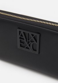 Svart lær lommebok med gull glidelås, teksturert overflate og en fremtredende svart logo-plakett som leser "ARMANI EXCHANGE."