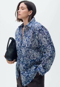 Camicia blu leggera a motivo, con design floreale paisley, maniche lunghe e chiusura con bottoni, abbinata a una borsa nera di tessuto.