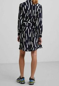 Donna che indossa un abito nero a maniche lunghe con motivi astratti bianchi, una cintura nera e scarpe sportive nere, blu e gialle, in piedi di spalle.
