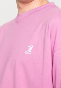 Vaaleanpunainen puuvillainen t-paita pyöreällä pääntiellä, jossa valkoinen kirjailtu kengurulogot vasemmalla rinnassa. Sileä rakenne, rento istuvuus.