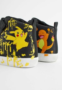 Schwarze Sneakers mit gelbem Spritzdesign, featuring Pikachu- und Charmander-Grafiken. Weiches Material, weiße Gummisohlen, Klettverschlüsse.
