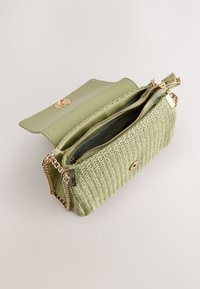Sac à main tissé vert clair avec une bandoulière chaîne dorée, rabat ouvert et compartiment intérieur zippé visible.