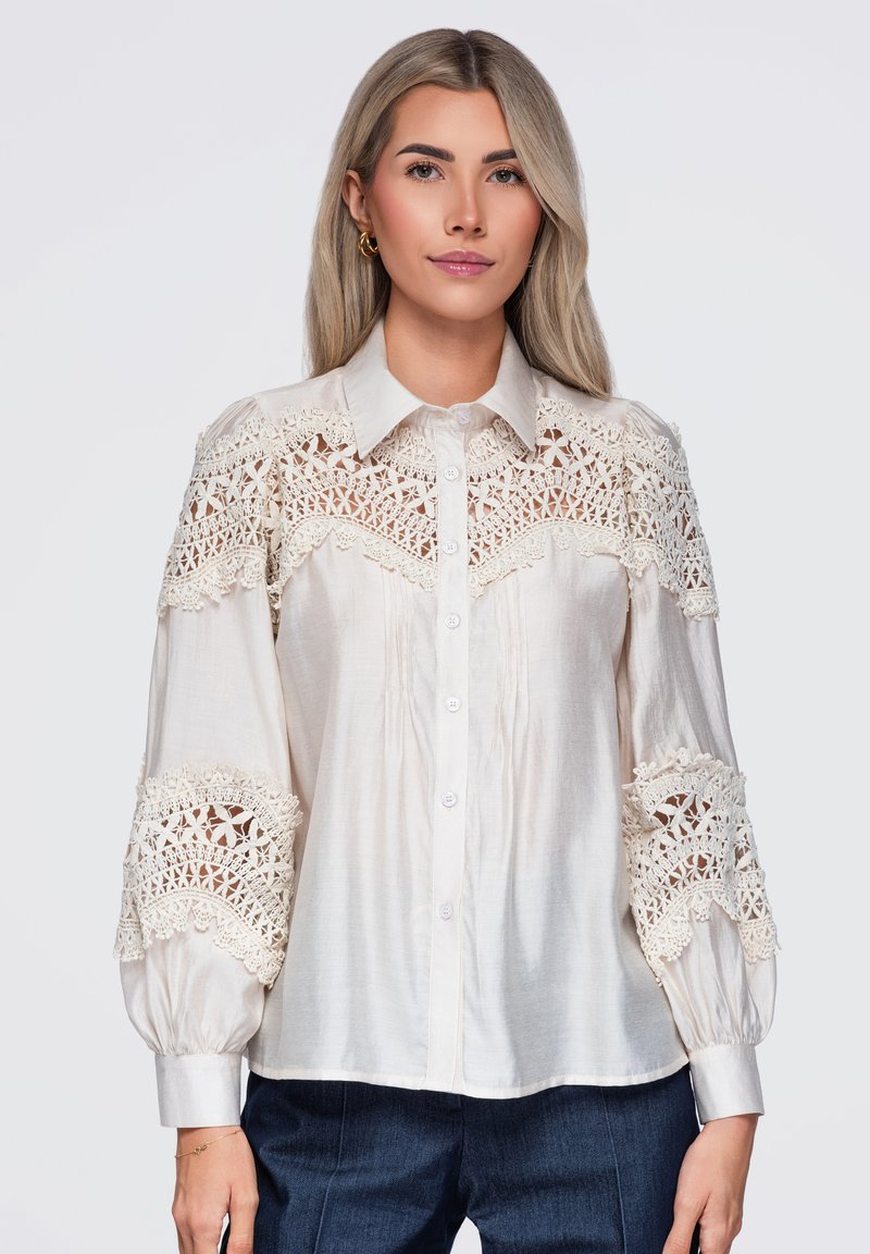 Femme aux longs cheveux blonds portant un chemisier blanc avec des détails en dentelle finement travaillés sur les épaules et les manches, associée à un pantalon foncé.