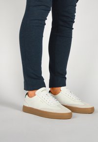 Baskets en cuir blanc avec des accents en suède gris clair, semelles en caoutchouc beige et lacets blancs, portées avec un pantalon ajusté bleu foncé.