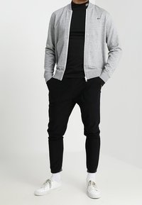 Šedá bomber bunda na zip přes černý rolák, doplněná černými tepláky a bílými teniskami se šněrováním a viditelnými podrážkami.