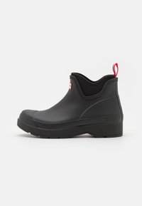 Bottine noire en caoutchouc avec un bout arrondi, semelle texturée, panneaux latéraux élastiques et une tirette rouge à l'arrière.