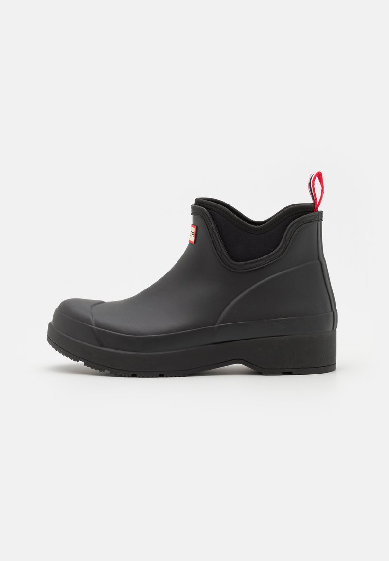 Bottine noire en caoutchouc avec un bout arrondi, semelle texturée, panneaux latéraux élastiques et une tirette rouge à l'arrière.