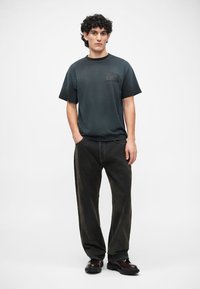 Donkergroene korte mouwen t-shirt met een bedrukte logo op de borst, gecombineerd met losse zwarte spijkerbroeken en zwarte schoenen.