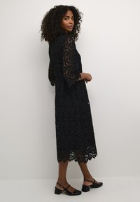 Black lace dress featuring a floral pattern, 3/4 length sleeves, and scalloped hem. Paired with black block-heel shoes.