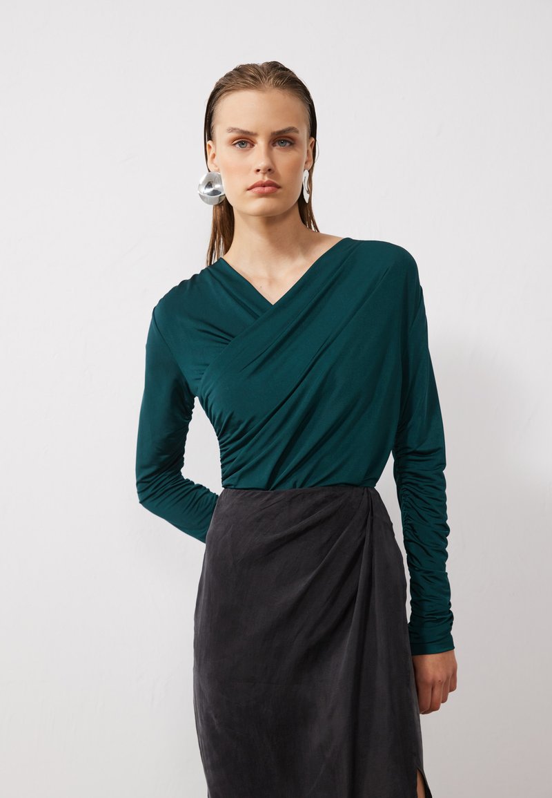 Teal long-sleeve top with a wrapped design, paired with a black draped skirt. The top features textured fabric and fitted sleeves.
