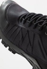 Chaussure de sport noire avec une tige en mesh texturé, un bout renforcé et une semelle en caoutchouc avec des crampons pour la traction. Les lacets sont noirs et plats.