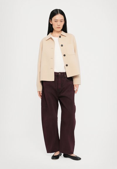 Chaqueta beige corta con tres botones negros, combinada con pantalones de pierna ancha burdeos oscuros. El modelo lleva una blusa blanca y zapatos planos negros.
