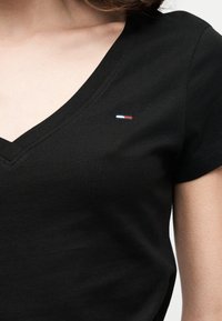 Μαύρο κοντομάνικο μπλουζάκι με V-λαιμόκοψη που φοριέται από άτομο, με μικρό ορθογώνιο λογότυπο με λευκές, κόκκινες και μπλε ρίγες στο αριστερό στήθος.