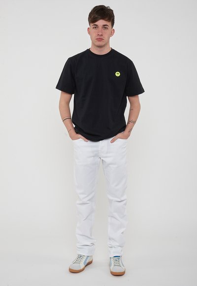 T-shirt en coton noir avec un petit logo jaune, associé à un jean en denim blanc et des baskets gris clair avec des accents bleus et beiges.