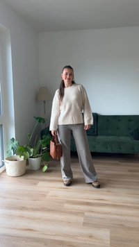 Cremefarbener Wollmischpullover mit weiten Ärmeln, graue Hose und braune Handtasche. Grünes Sofa und Pflanzen in einem hellen Innenraum.
