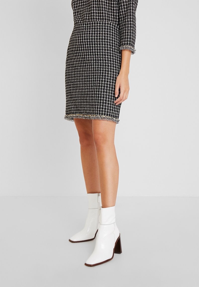 Femme portant un tailleur jupe à carreaux noir et blanc avec des ourlets à franges et des bottines blanches à talons, debout sur un fond uni.