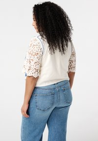 Witte top met haakwerk mouwen en blauwe stiksels, gecombineerd met lichtblauwe high-waisted jeans. De stof is zacht en textuur. 
