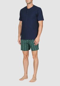Maglietta henley a maniche corte blu navy con tre bottoni, abbinata a pantaloni corti verdi con motivi di barche bianche e vita elastica.
