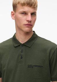 Olīvu zaļš polo krekls, kas izgatavots no tekstūras auduma, ar trim melniem pogām un tumšu izšūtu logotipu. Īsas piedurknes un klasiskā apkakles dizains.