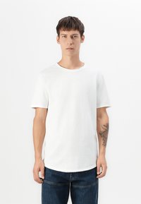 Weißes Baumwoll-T-Shirt mit kurzen Ärmeln, Rundhalsausschnitt und lockerer Passform. Hat einen leicht abgerundeten Saum. Getragen mit dunkelblauen Denim-Jeans.