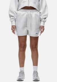 Pantaloni corti bianchi in cotone con vita elastica e coulisse. Presentano dettagli ricamati in blu. Indossati con scarpe sportive di colore chiaro.