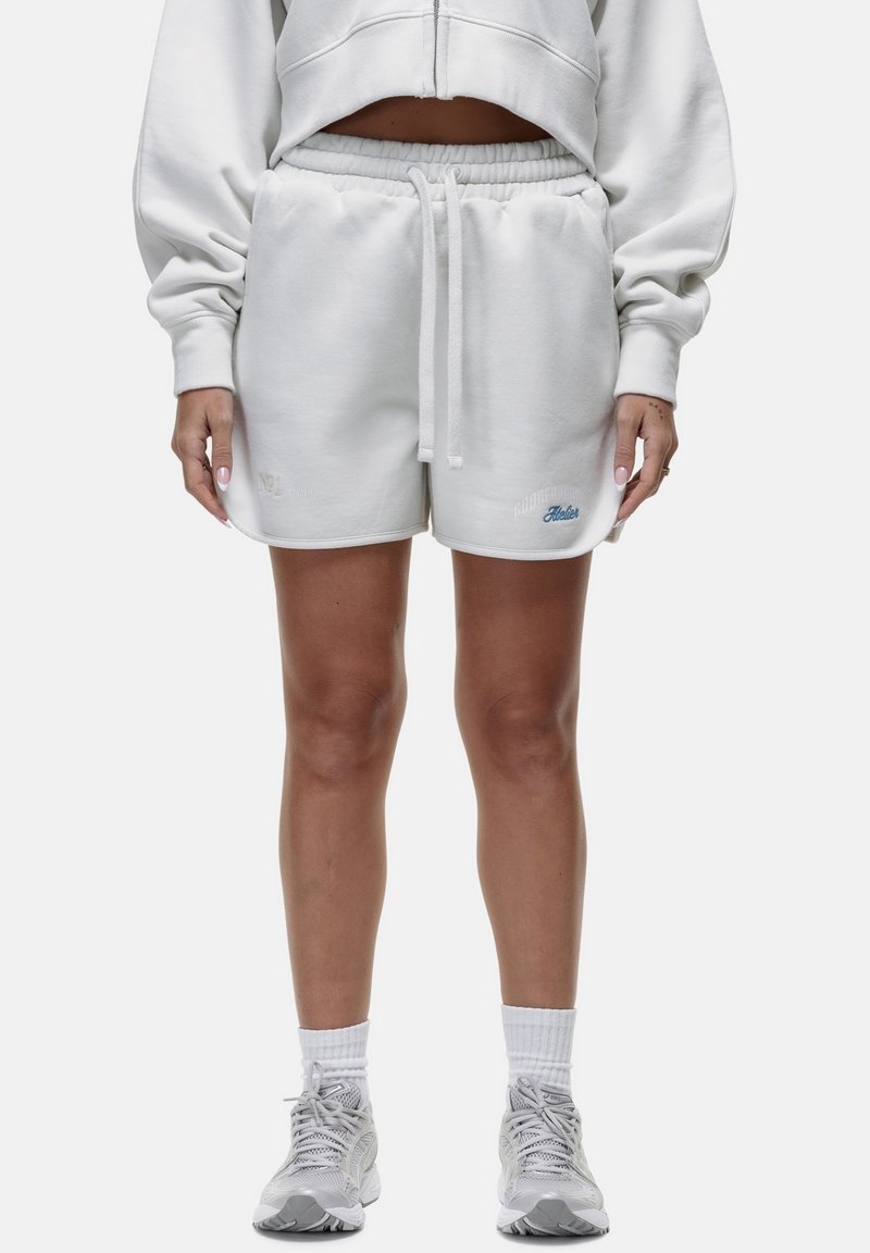 Pantaloni corti bianchi in cotone con vita elastica e coulisse. Presentano dettagli ricamati in blu. Indossati con scarpe sportive di colore chiaro.