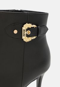 Μαύρη δερμάτινη μποτάκι μέχρι τον αστράγαλο με χρυσό διακοσμητικό κούμπωμα, με λείο υλικό, μυτερή μύτη και ψηλό τακούνι. Παρουσιάζει ραφές με λεπτομέρεια.