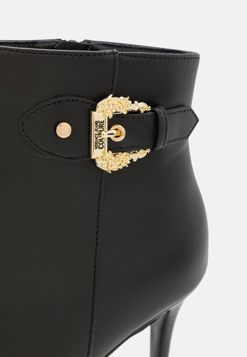 Stivale alla caviglia in pelle nera con una fibbia decorativa dorata, caratterizzato da una texture liscia, punta affusolata e tacco alto. Presenti dettagli cuciti.