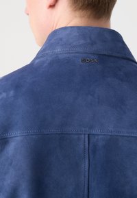 Ναυτικό μπλε σουέντ μπουφάν με γιακά, που διαθέτει μια διακριτική γυαλάδα, ραφές στην κορυφή και μια μαύρη λεπτομέρεια λογότυπου "BOSS" κοντά στον λαιμό.